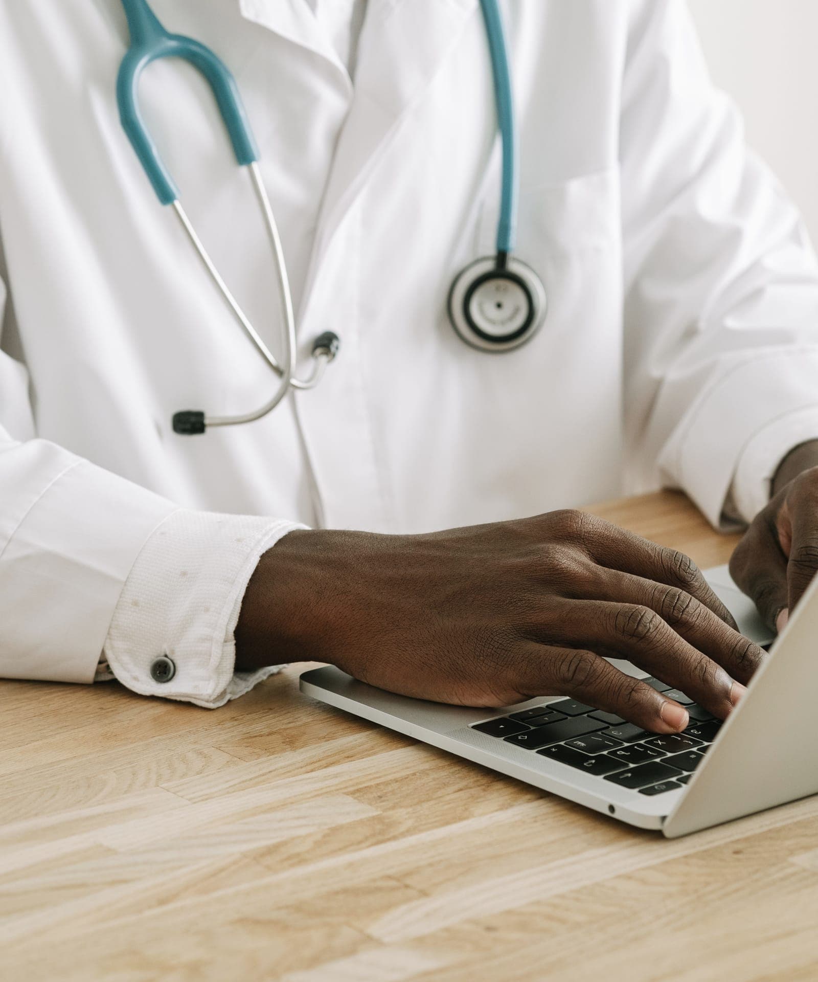 A clinician in a white coat with a teal stethoscope typing on a laptop at a wooden desk.