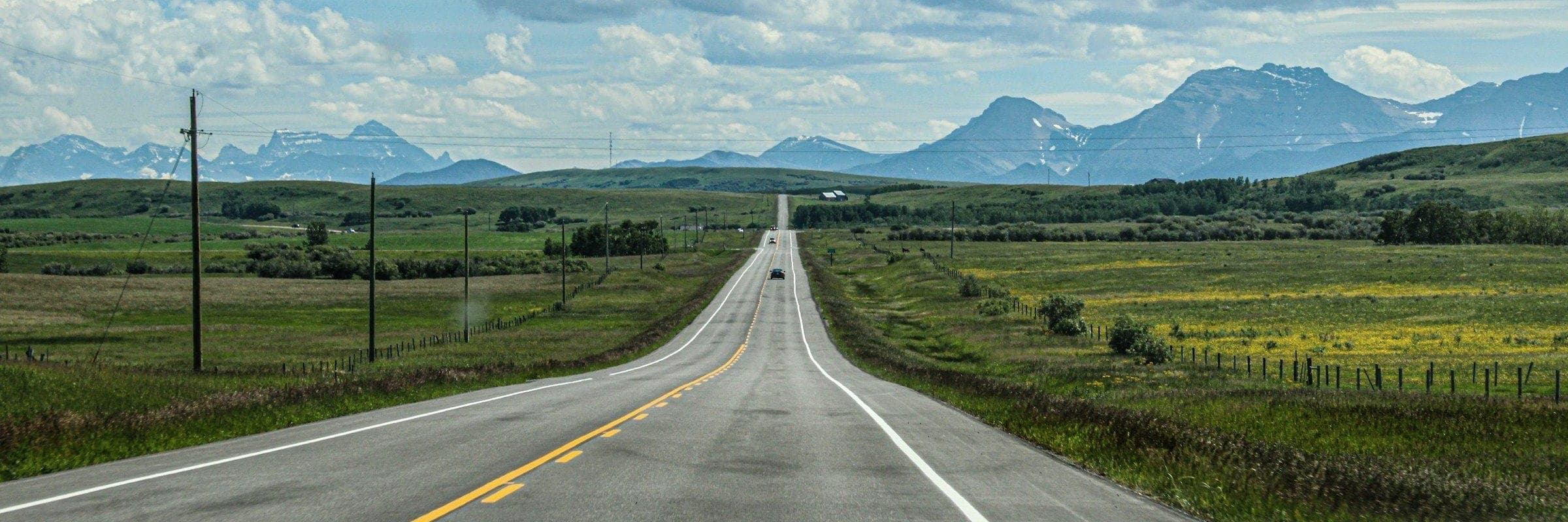 An empty two-lane highway running toward distant mountains under a wide, cloudy sky.